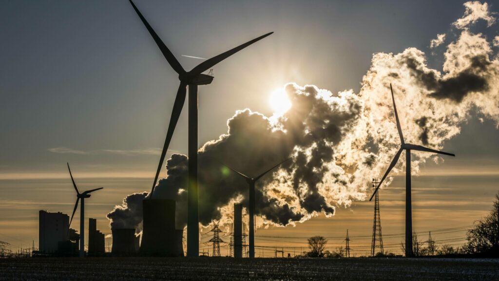 Windräder, Kraftwerke und Stromleitungen als Silhouetten vor Himmel und Sonne.