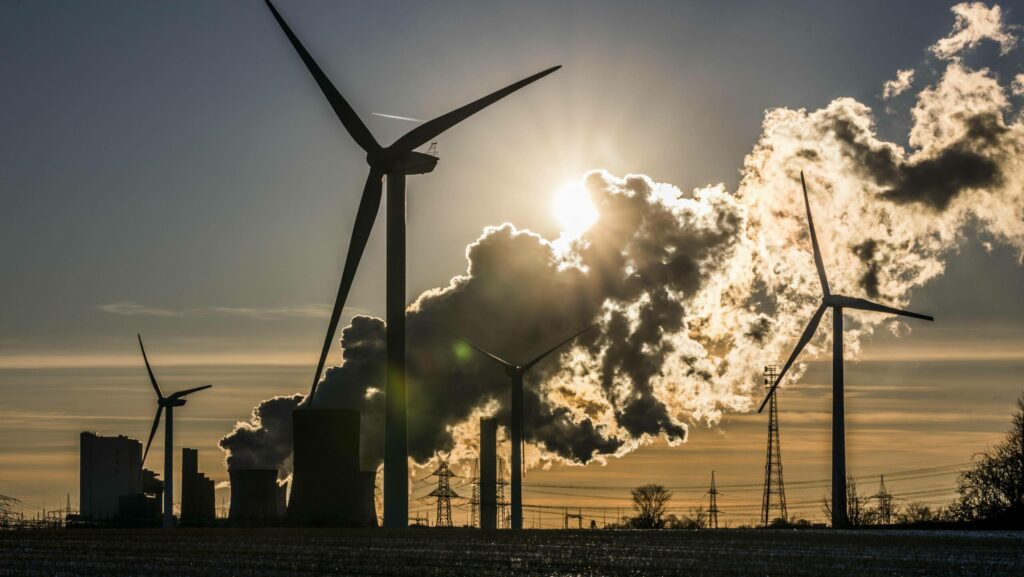 Windräder, Kraftwerke und Stromleitungen als Silhouetten vor Himmel und Sonne.