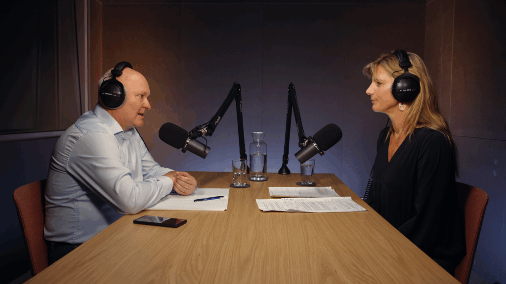 Zwei Personen sitzen an einem Holztisch mit Kopfhörern und Mikros in einem Podcast-Studio und unterhalten sich.