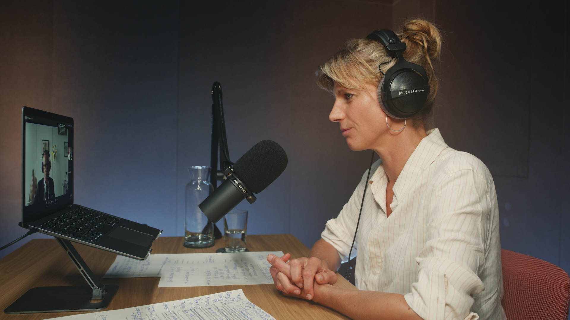 Eine Person sitzt an einem Holztisch mit Kopfhörern und Mikros in einem Podcast-Studio und schaut auf einem Laptop, wo der Gast über Video zugeschaltet ist.