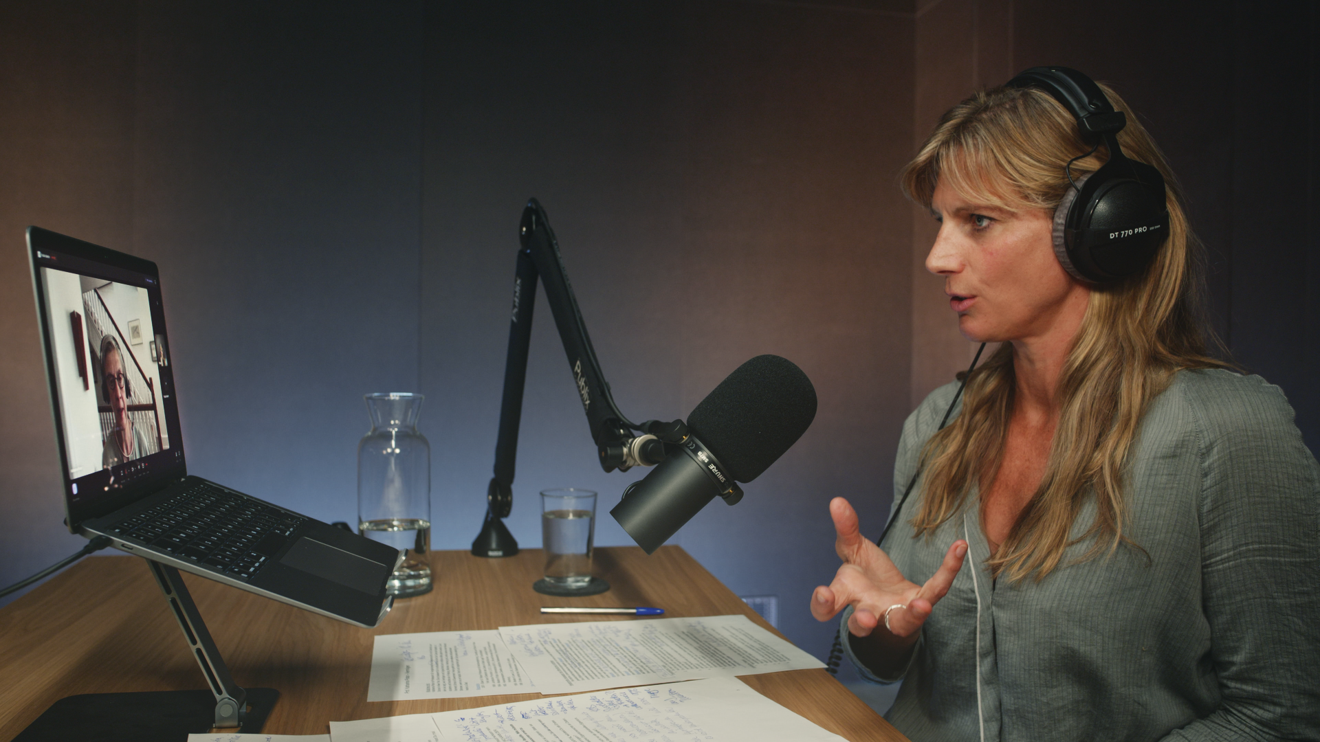 Eine Person sitzt an einem Holztisch mit Kopfhörern und Mikros in einem Podcast-Studio und spricht mit Handgeste in Richtung eines Laptop, wo der Gast über Video zugeschaltet ist.
