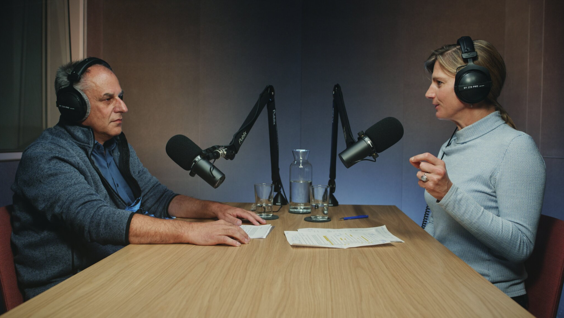 Zwei Person sitzen sich gegenüber an einem Holztisch mit Kopfhörern und Mikros in einem Podcast-Studio. Die Person rechts spricht und gestikuliert, die links hört aufmerksam zu.
