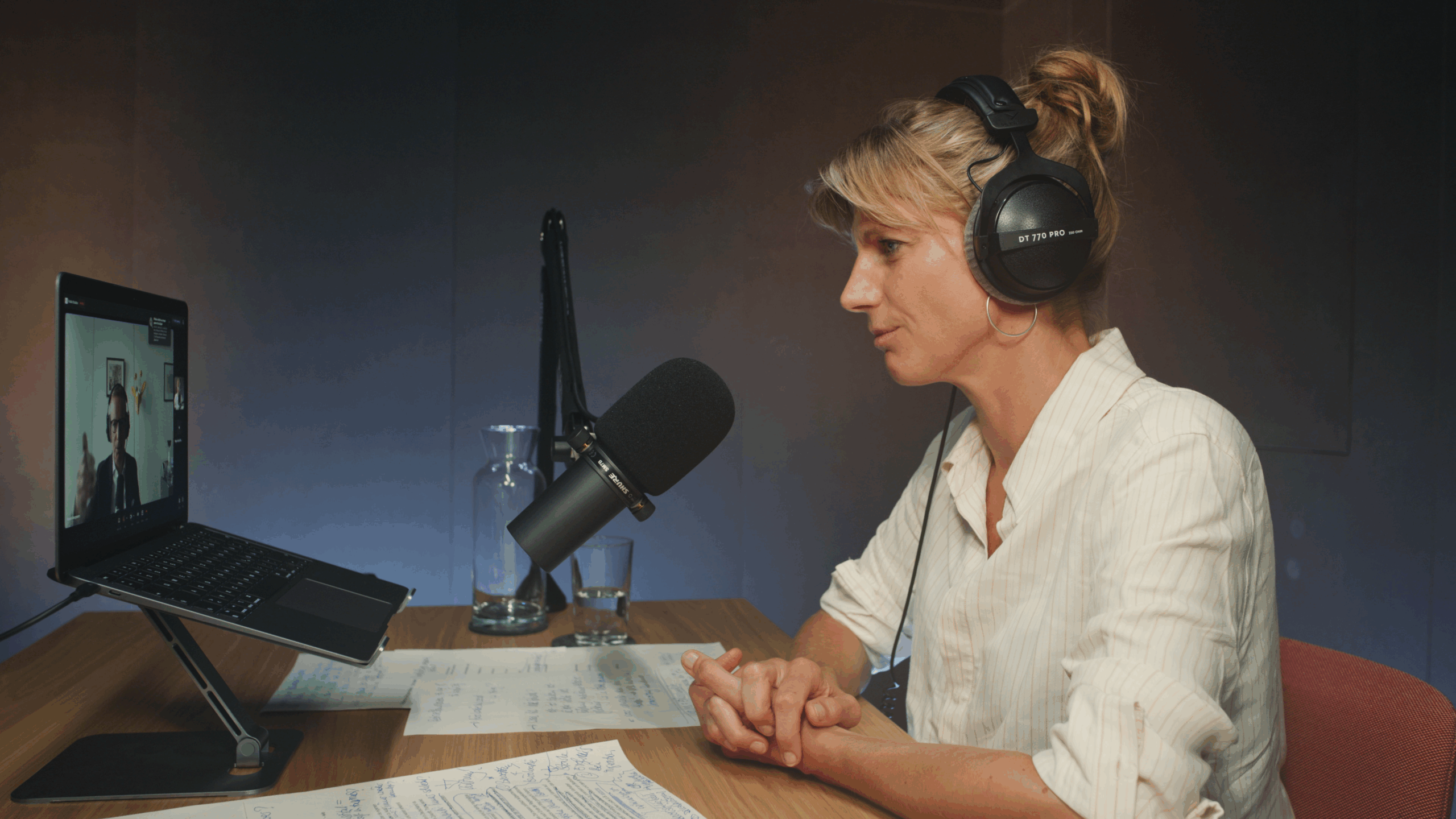 Eine Person in weißer Bluse mit Kopfhörern über den blonden Haaren sitzt von einem Studiomikrofon und schaut auf einen Laptop.