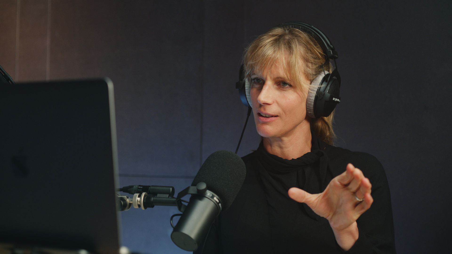Eine Person sitzt mit Kopfhörer vor einem Mikrofon und spricht mit Geste in Richtung eines Laptops.