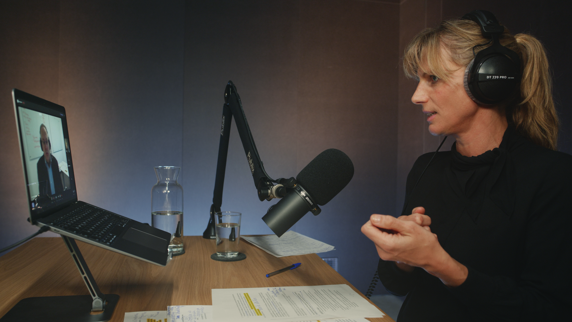 Eine Person sitzt an einem Holztisch mit Kopfhörern und Mikro in einem Podcast-Studio und spricht mit Handgeste in Richtung eines Laptop, wo der Gast über Video zugeschaltet ist.