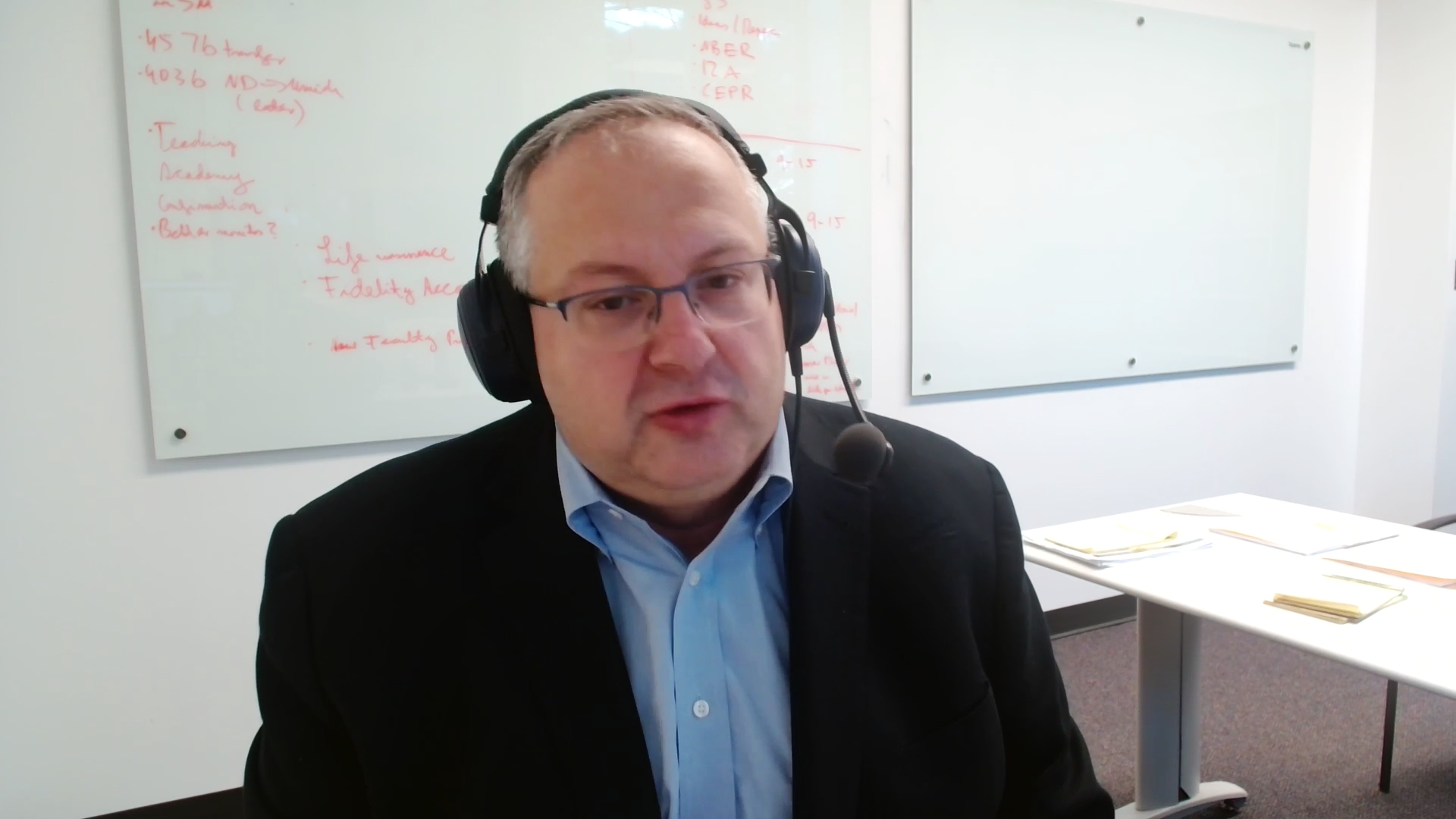 Eine Person sitzt mit Headset vor einem Whiteboard und spricht in die Kamera.