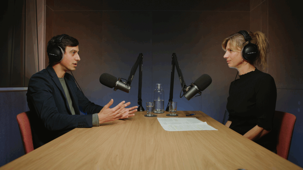 Zwei Person sitzen sich gegenüber an einem Holztisch mit Kopfhörern und Mikros in einem Podcast-Studio. Die Person links spricht und gestikuliert.
