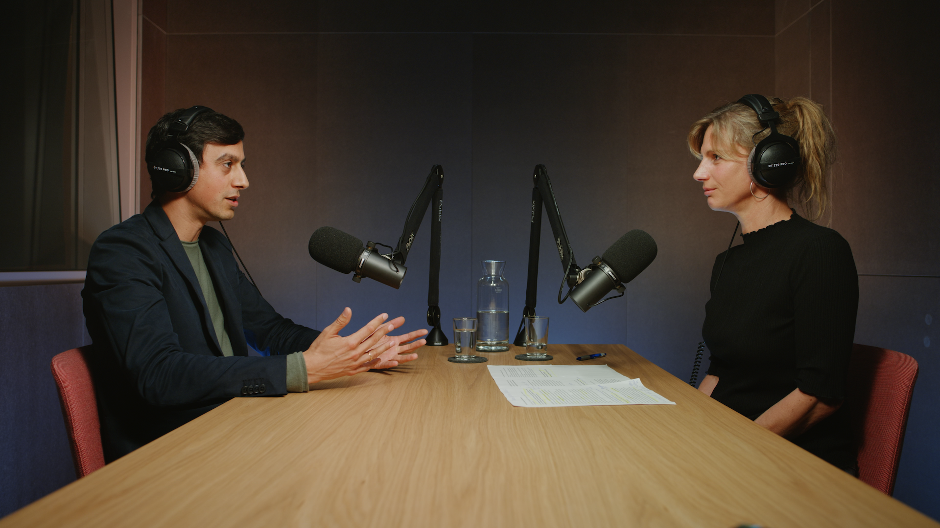 Zwei Person sitzen sich gegenüber an einem Holztisch mit Kopfhörern und Mikros in einem Podcast-Studio. Die Person links spricht und gestikuliert.