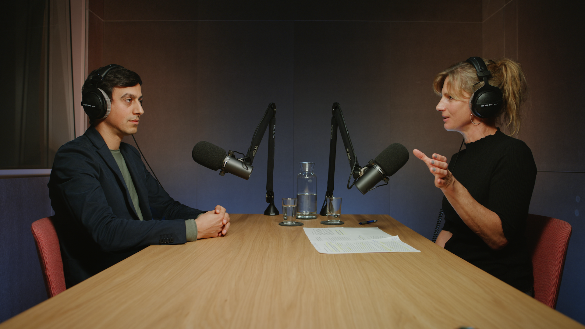 Zwei Personen sitzen sich gegenüber an einem Holztisch mit Kopfhörern und Mikros in einem Podcast-Studio. Die Person rechts spricht und gestikuliert.
