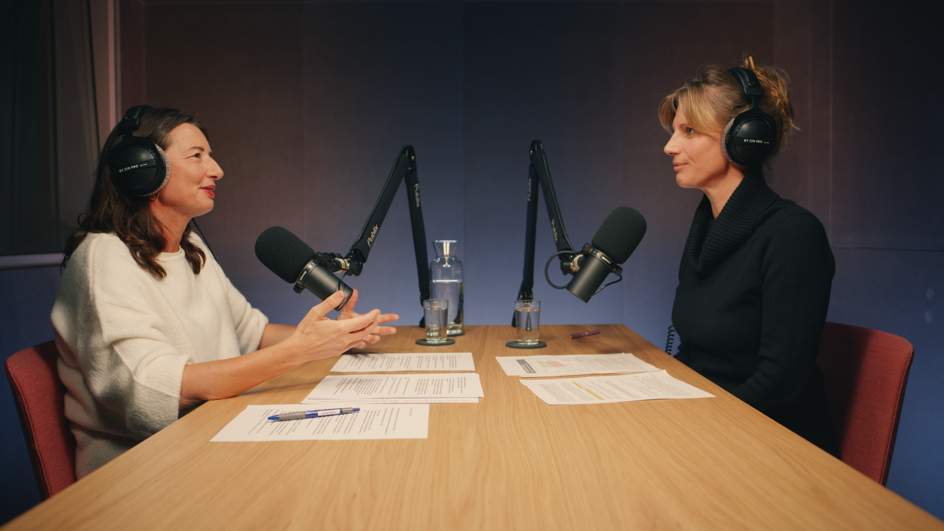 Zwei Personen sitzen an einem Holztisch mit Kopfhörern und Mikros in einem Podcast-Studio und unterhalten sich.