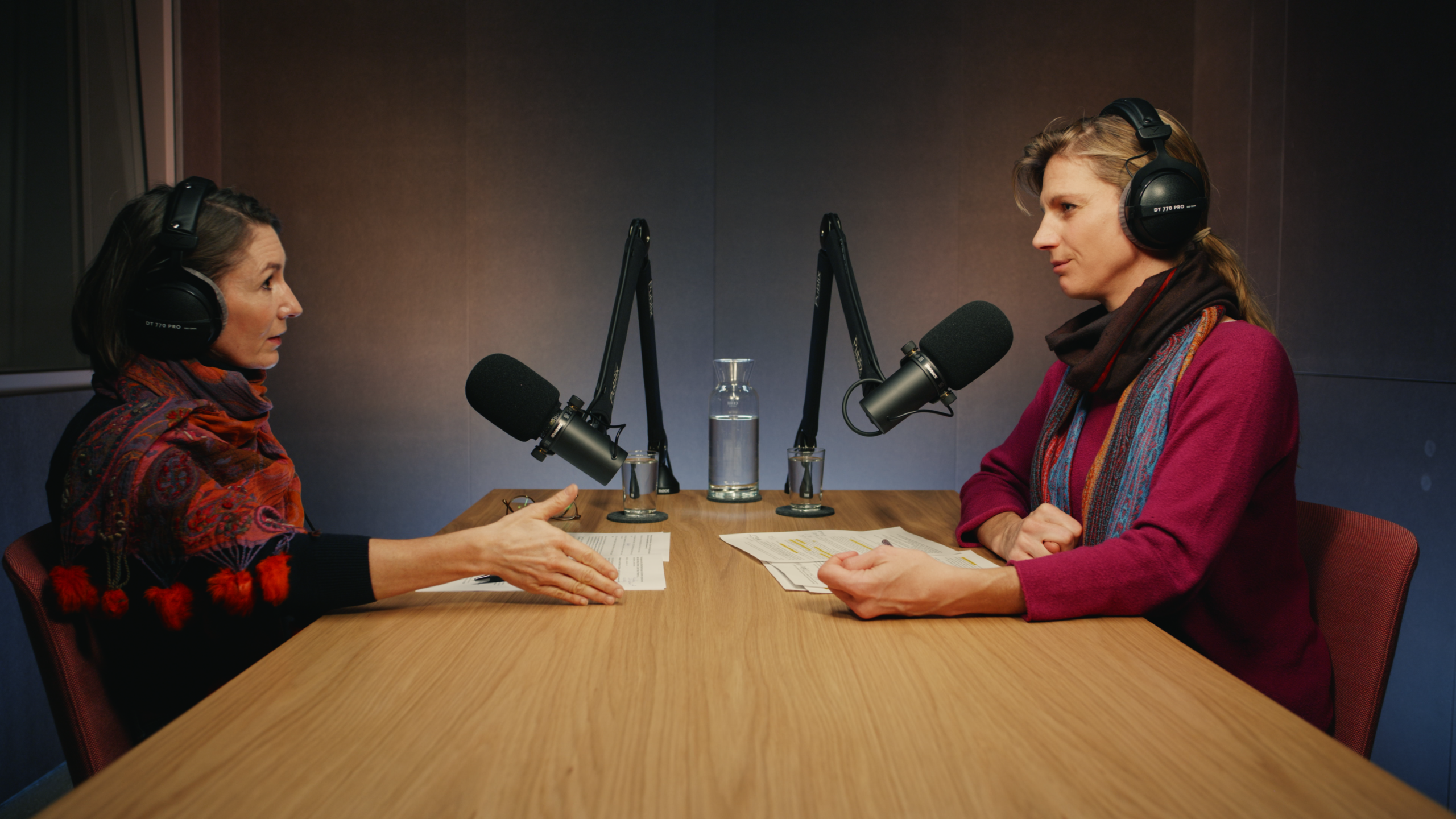 Zwei Personen sitzen an einem Holztisch mit Kopfhörern und Mikros in einem Podcast-Studio und unterhalten sich.