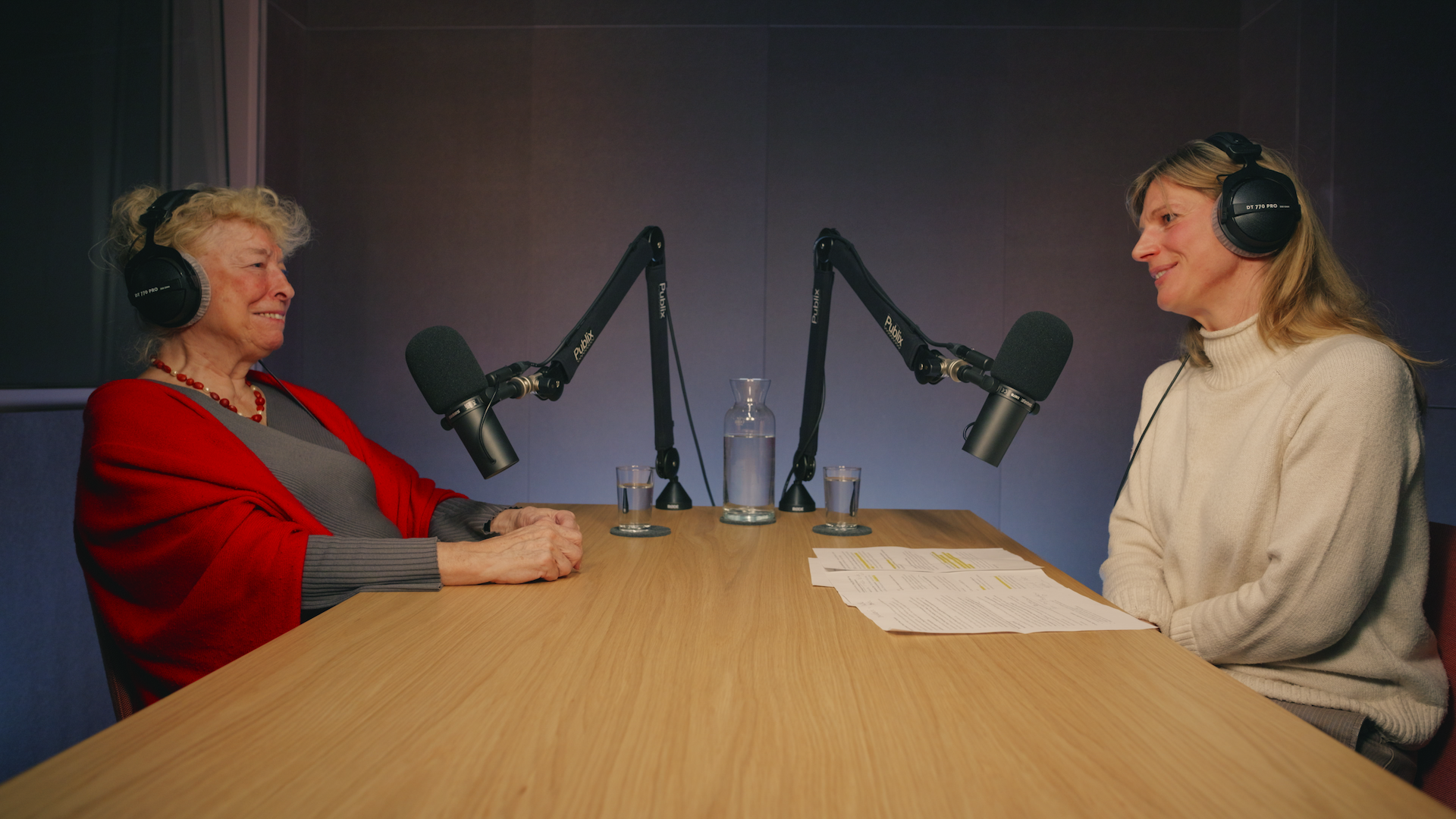 Zwei Personen sitzen vor Mikros an einem Holztisch.