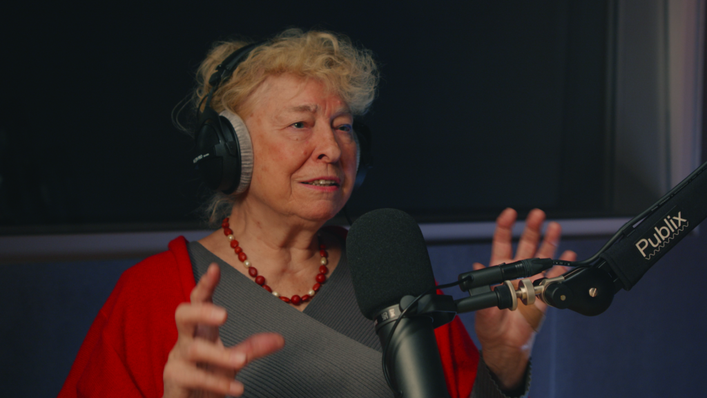 Eine Person mit blonden Haaren und roter Jacke mit Kopfhörern vor einem Mikro