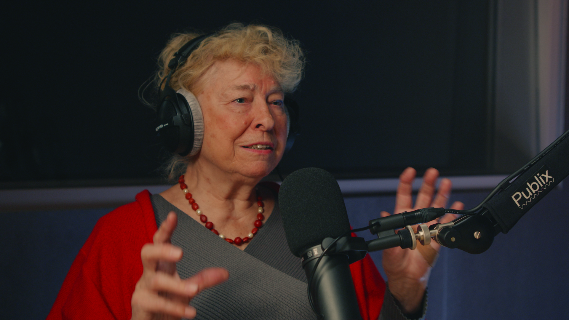 Eine Person mit blonden Haaren und roter Jacke mit Kopfhörern vor einem Mikro