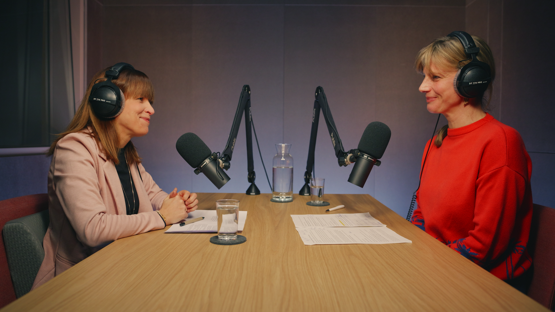 Zwei Personen sitzen vor Mikros an einem Holztisch im Studio und lächeln sich an.