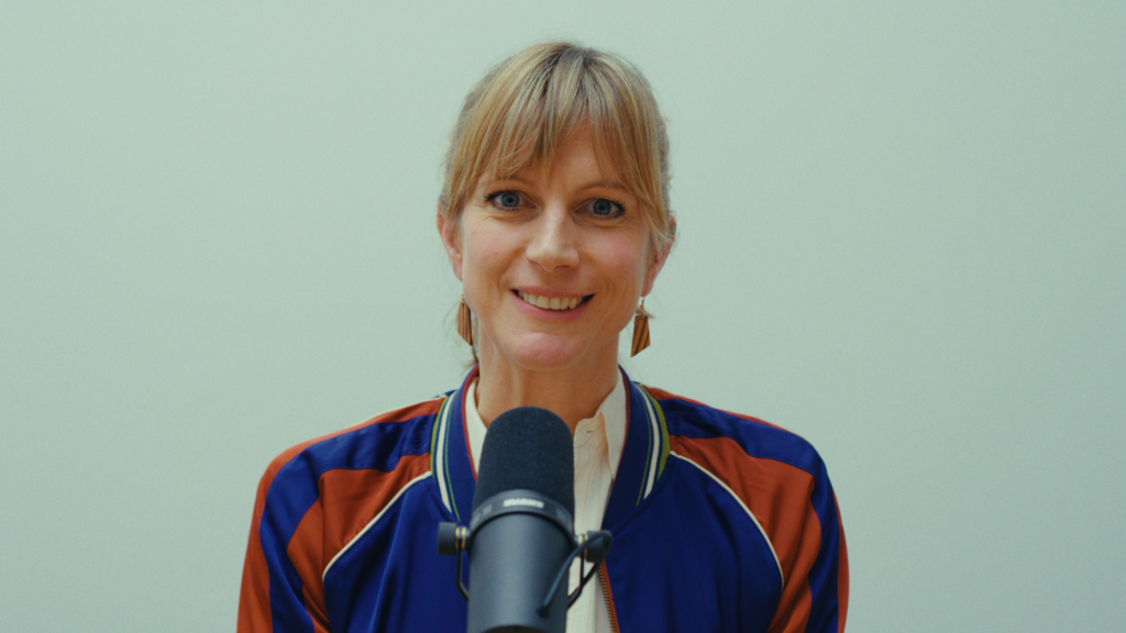 Eine Person mit blonden Haaren, Pony und Jacke mit bunten Aufnähern lächelt in die Kamera. Vor ihr steht ein Mikrofon.