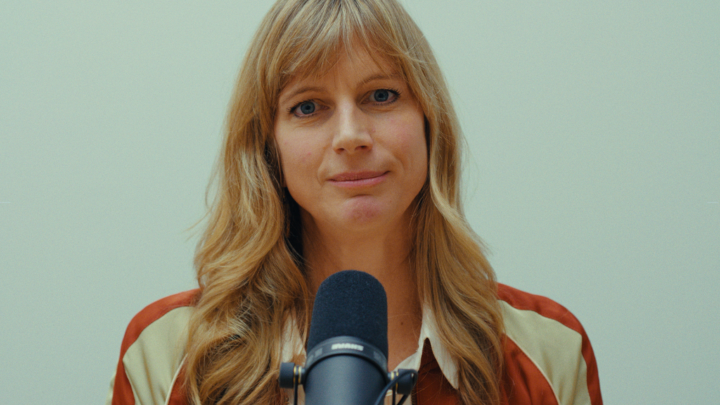 Eine Person mit langen blonden Haaren schaut optimistisch in die Kamera, vor ihr ein Mikrofon.