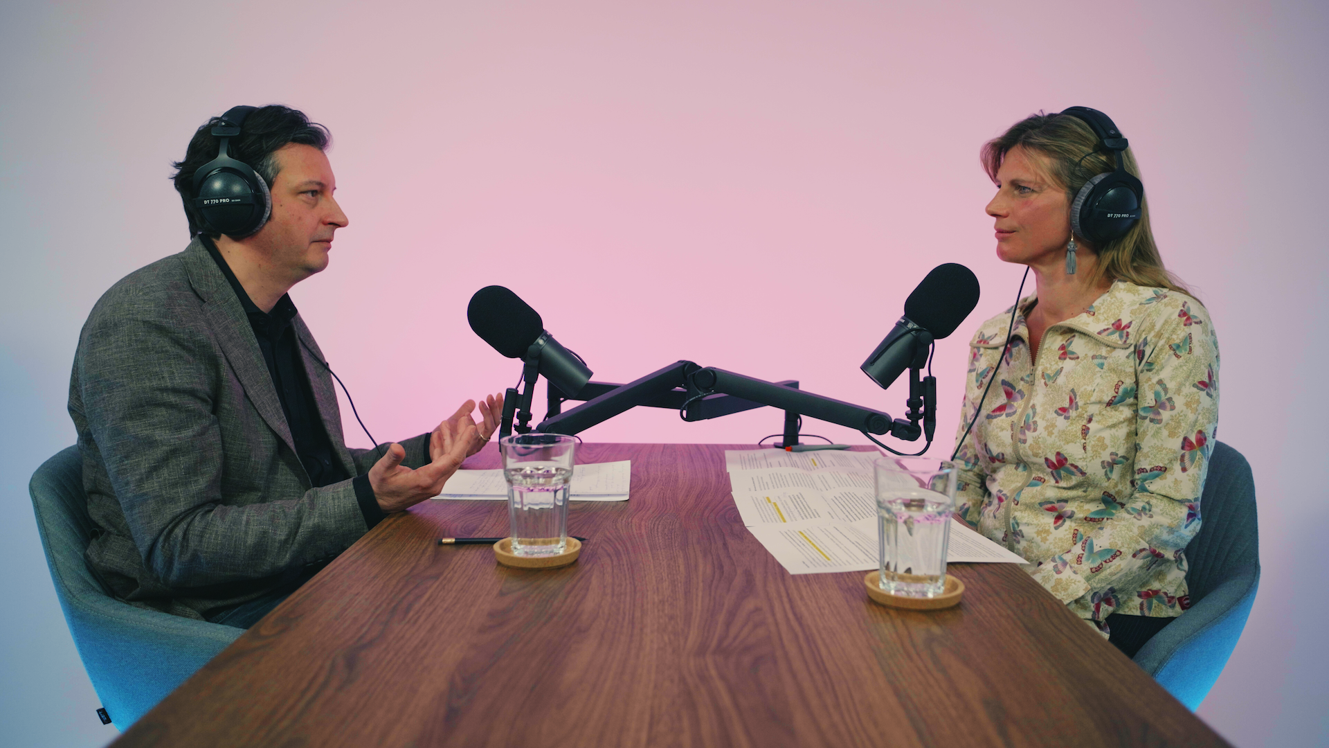 Zwei Personen sitzen sich gegenüber an einem Holztisch mit Kopfhörern und Mikros in einem Podcast-Studio. Die Person links spricht und gestikuliert.