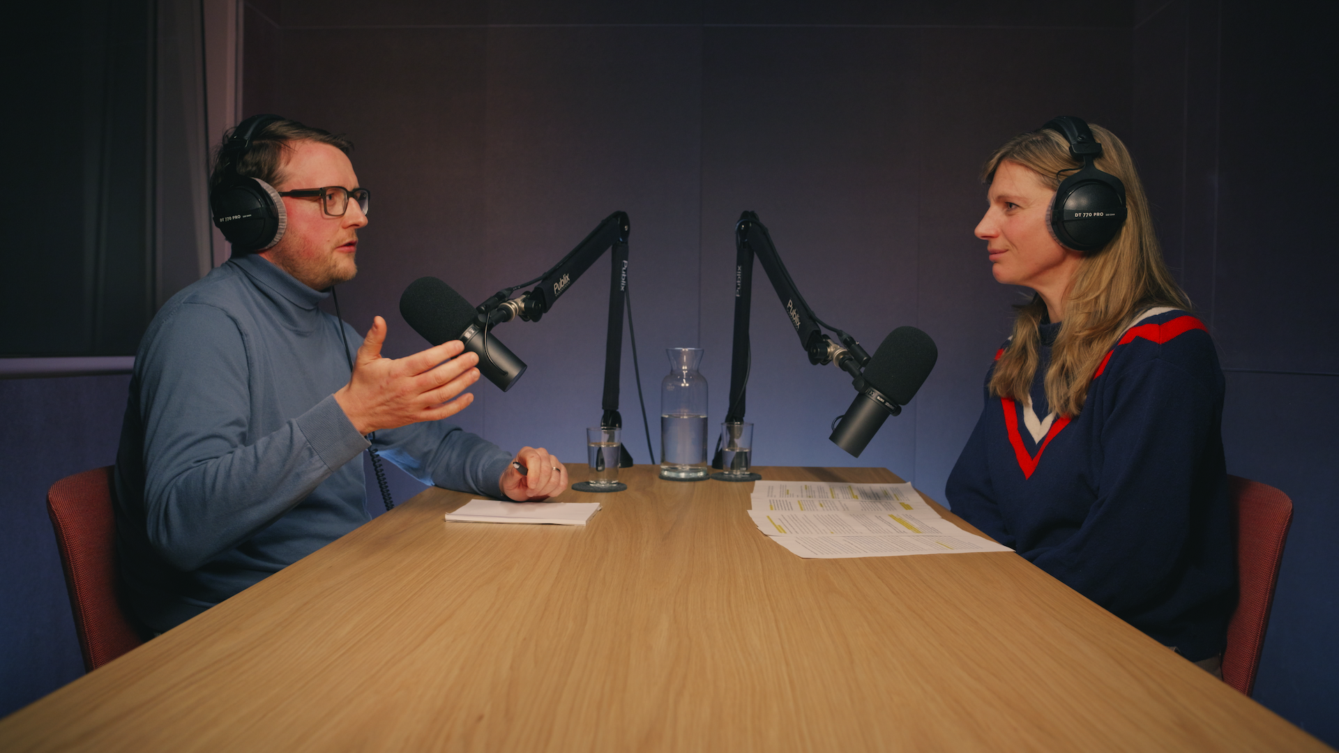 Zwei Personen sitzen sich gegenüber an einem Holztisch mit Kopfhörern und Mikros in einem Podcast-Studio. Die Person links spricht und gestikuliert.