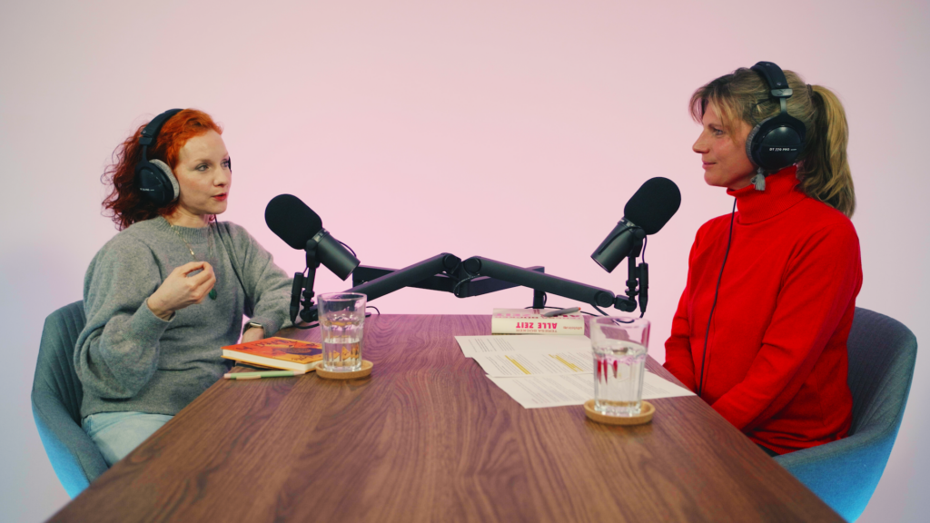 Zwei Personen sitzen sich gegenüber an einem Holztisch mit Kopfhörern und Mikros in einem Podcast-Studio. Die Person links spricht und gestikuliert.