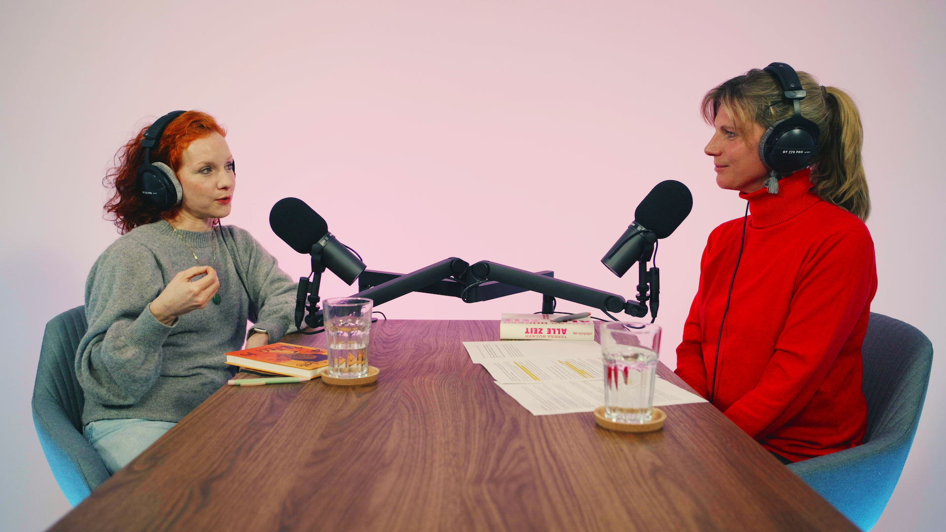 Zwei Personen sitzen sich gegenüber an einem Holztisch mit Kopfhörern und Mikros in einem Podcast-Studio. Die Person links spricht und gestikuliert.