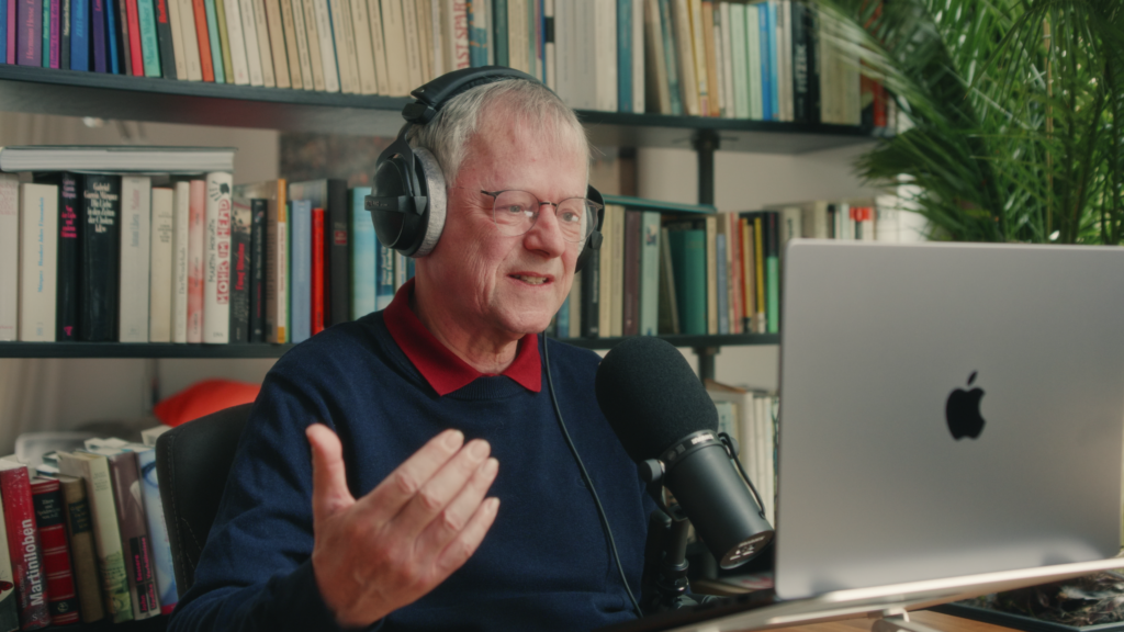 Ein Mann vor einem Bücherregal spricht in ein Mikrofon und blickt in eine Laptop-Kamera.