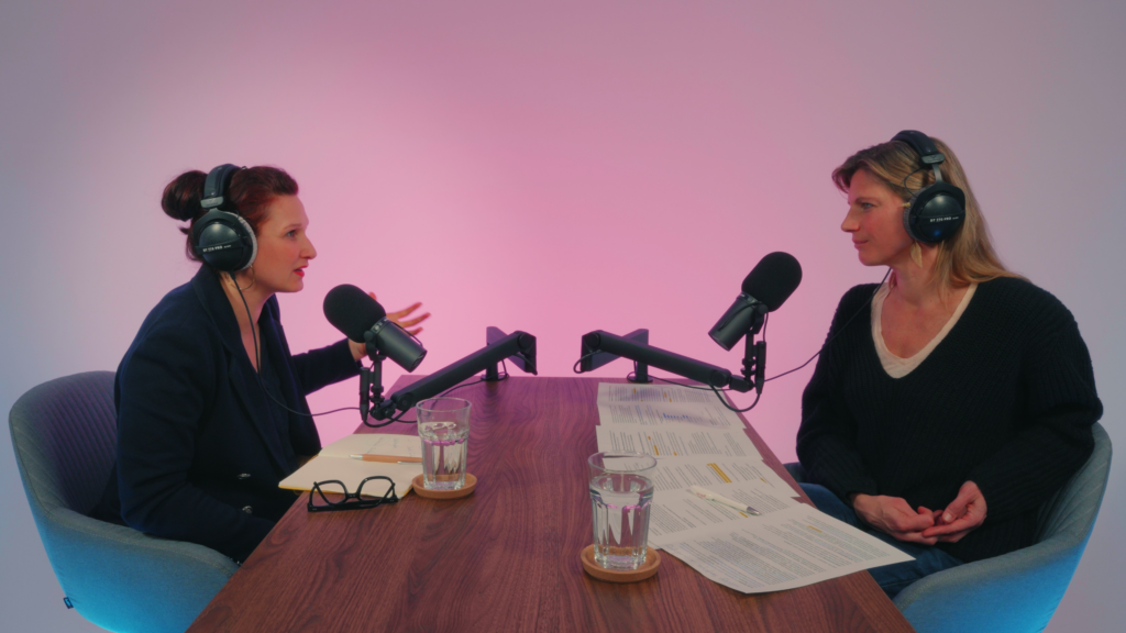Zwei Personen sitzen sich gegenüber an einem Holztisch mit Kopfhörern und Mikros in einem Podcast-Studio. Die Person rechts/links spricht und gestikuliert.