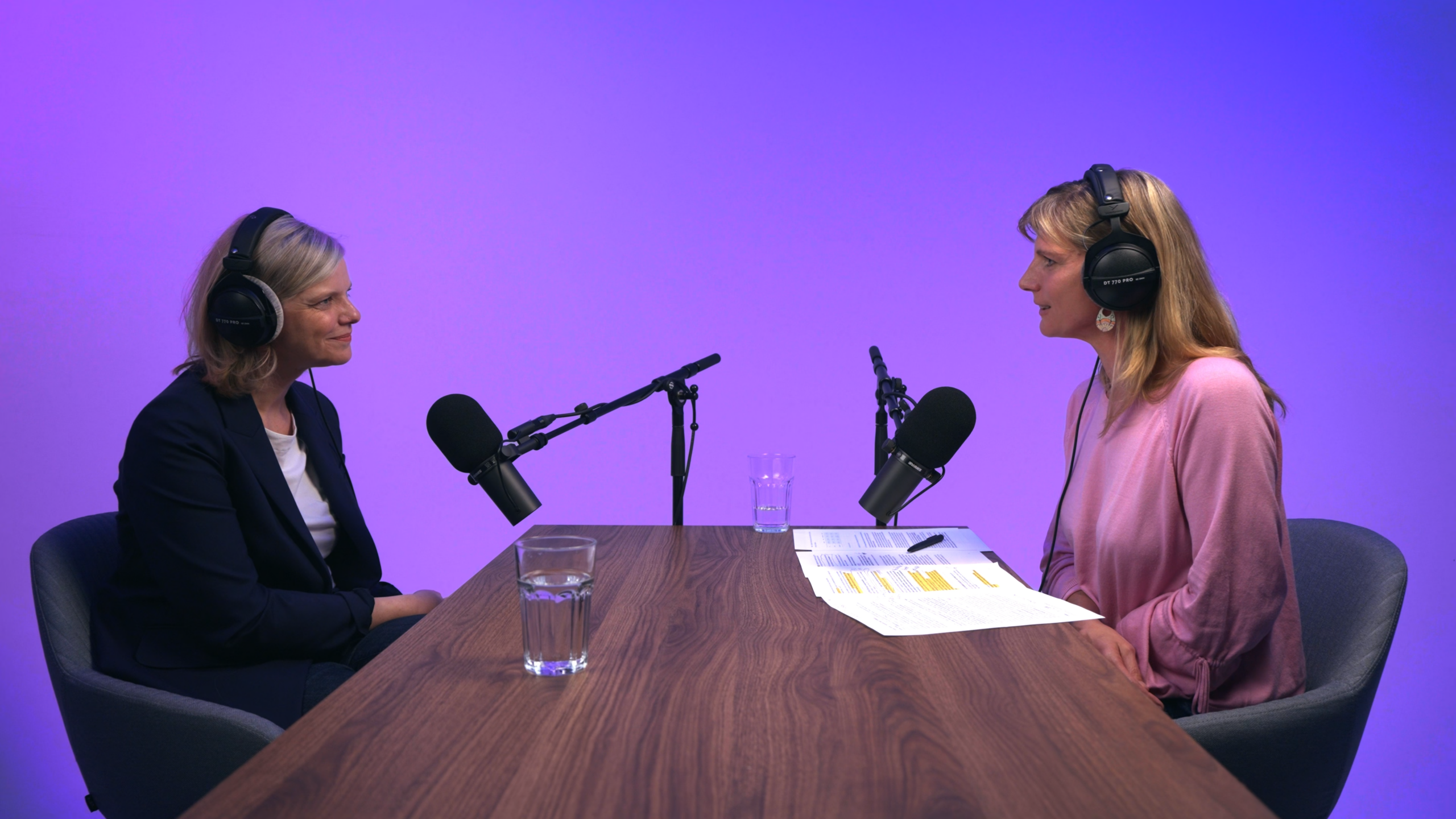 Zwei Frauen mit Kopfhörern sitzen sich an einem Holztisch bei einer Podcast-Aufnahme vor violettem Studiohintergrund gegenüber.
