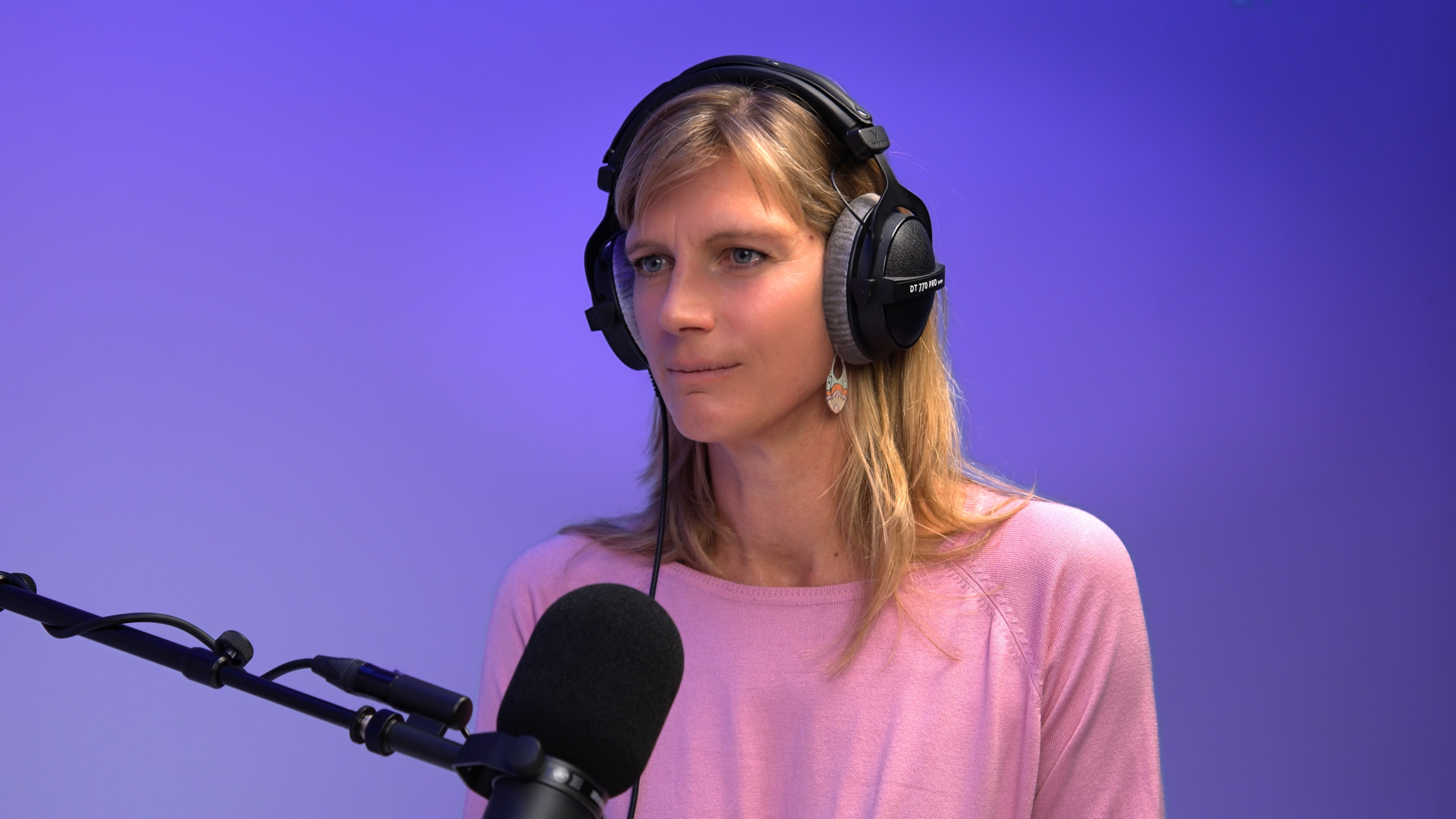 Maja Goepel mit Over-Ear-Kopfhörern und rosa Oberteil vor einem Mikrofon in einem Studio mit violett-blauem Hintergrund.